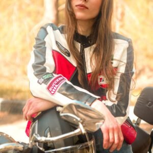 Woman wearing a biker jacket leaning on a motorcycle, outdoors, captured in warm daylight.